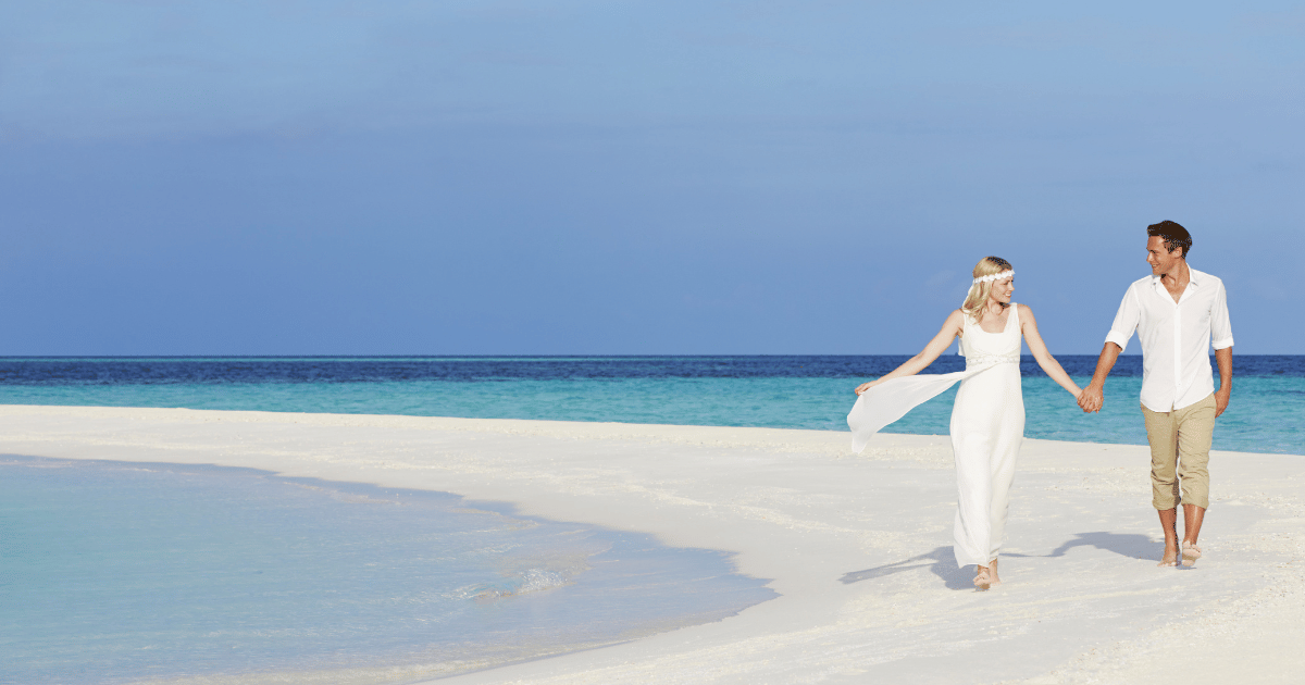 白い砂浜を手をつないで歩くカップル。背景には青い海と空が広がる